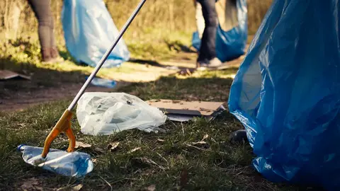 Volunteer using tongs tool to collect trash and plastic waste from the woods, storing rubbish in a garbage bag and clean the natural environment. Activist grabbing litter. Close up. Camera B.